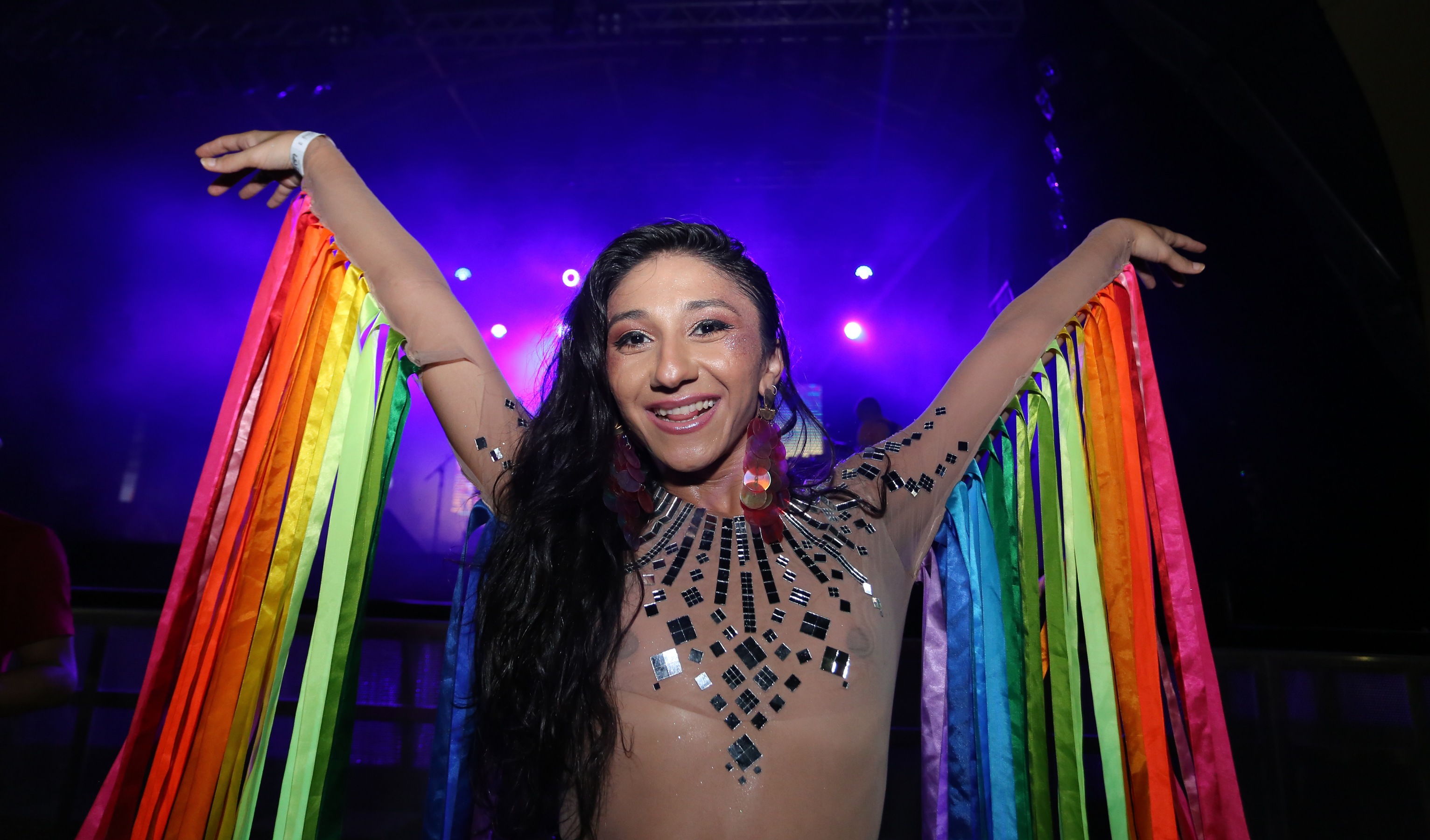 mulher fantasiada com fitas coloridas nos braços levantados e sorrindo para a foto